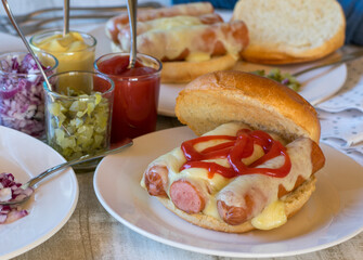Hot dog with cheese and ketchup on a burger bun served on a kitchen table with pickles, red onions, mustard and ketchup jar