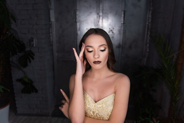 Fototapeta na wymiar Close up beauty portrait of girl, holds her hand to head, headache. Brunette with brown eyes, professional evening make-up, dark lipstick, smoky eyes in gold corset. Festive, party. Silver background
