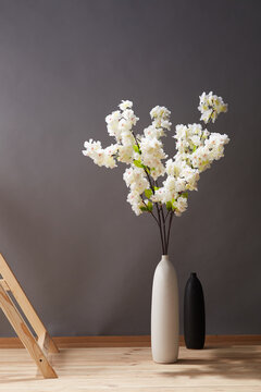 Still life: flowering branches of white sakura in white vase near wooden ladder and empty black vase on the wooden floor. The splendid bouquet is outlined against gray wall. 