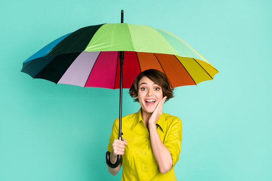 Photo portrait of amazed smiling girl touching cheek keeping parasol isolated on vibrant teal color background