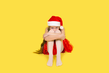 Sad teen girl in red dress and Santa Claus red hat sits and hug her knees. Girl in Santa hat isolated on yellow background