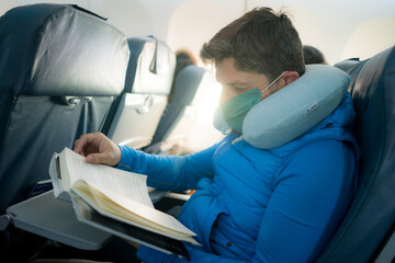 travel and tourism in times of covid19 - lifestyle portrait of young attractive Caucasian man in face mask and neck pillow reading book in airplane during flight
