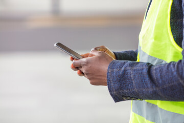 Foreman sending instructions via smartphone. Worker in vest standing inside of building in construction process, typing a message on smart phone and taking a break from work.