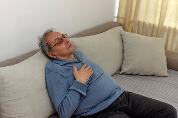 Fototapeta premium Worried senior man feeling unwell and having chest pain in the living room. Elderly man suffering from heart attack at home. Mature man feeling chest pain while sitting in the living room at home.