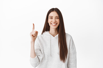 Cheerful smiling girl with perfect smile and clean face, pointing finger up at top logo, showing advertisement banner above, standing in casual hoodie, white background