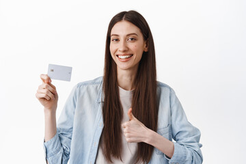 Cheerful brunette girl show credit card and thumbs up, recommending bank, praise contactless easy payment, good discounts, standing over white background