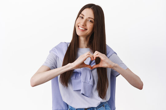 Like it. Smiling cute young woman showing heart gesture, tilt head and look adorable, I love you sign, standing against white background