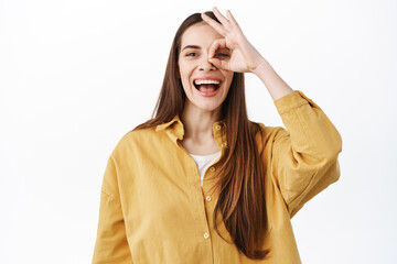 Happy laughing woman show ok sign and smiles, make okay gesture over eye and looking through, praise something good, excellent job, nice work, well done gest, standing over white background