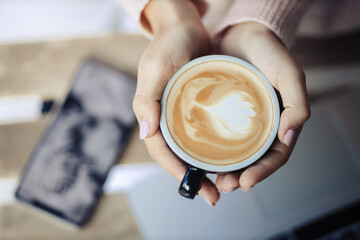 A girl with a beautiful delicate pink manicure holds a black cup with a fragrant cappuccino covered in foam. A woman works with a laptop and drinks coffee. 