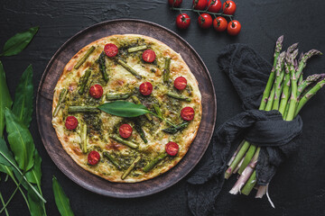 Homemade tarte flambee with green asparagus, tomatoes and wild garlic pesto on a wooden plate, top...