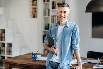 Portrait mature confident intelligent gray-haired caucasian man in casual stylish clothes, business man or manager, stands near the table, holds a laptop in hand, looking at camera, smiling friendly