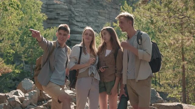 Tracking shot of group of young people with backpack standing on hiking trail in woods and looking at map on their mobile phone. They are discussing directions and laughing