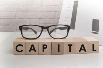 The word CAPITAL is written on wooden cubes standing on the white surface of the table, the GLASSES are on the cubes