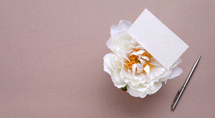 White empty paper card, white peony and pen on the beige surface.Empty space