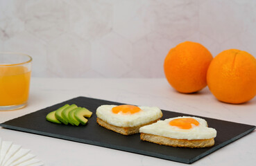 Breakfast.Heart shaped fried egg sandwiches with avocado slices on a stone plate and orange juice.
