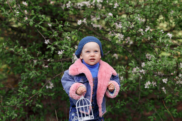 a small child in a blue knitted hat, blue and pink jacket stands near a flowering tree and looks up