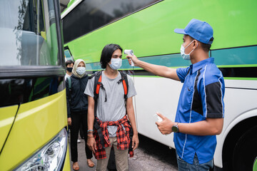 A bus crew in blue uniforms and a hat using a thermo gun inspects the male passenger in the mask before boarding the bus