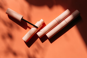 open tube with eyeliner brush next to closed pink tubes with mascara and liquid lipstick, lip gloss on a peach background with shadows