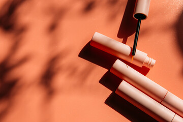 open tube with eyeliner brush next to closed pink tubes with mascara and liquid lipstick, lip gloss on a peach background with shadows