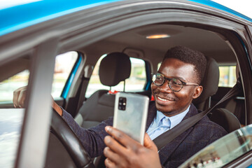 Handsome young businessman looking on his smart phone while sitting on the front seat and driving a car. Businessman Dials a Message on the Smartphone in the Car. Transport, Business Trip