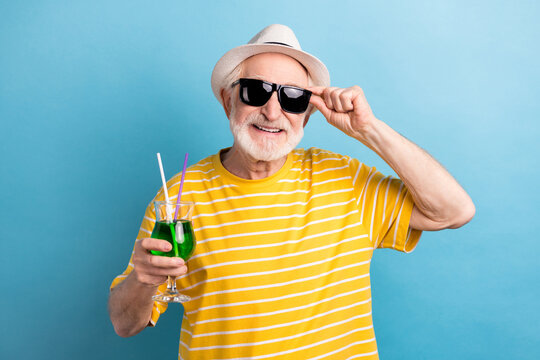 Photo of funny cool age gentleman wear yellow t-shirt cap dark spectacles smiling holding cocktail isolated blue color background