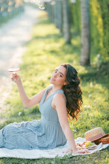 Naklejka premium Beautiful young woman with curly hair drinks pink champagne in the blooming sakura garden. Picnic in nature.