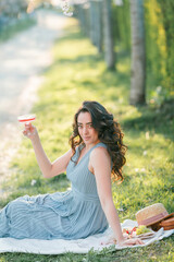 Naklejka premium Beautiful young woman with curly hair drinks pink champagne in the blooming sakura garden. Picnic in nature.