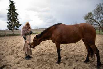 Eine blonde Frau aus den Mittelalten Zeiten kuschelt mit ihrem Pferd. Sie hat Fell auf den Schultern, ein Rock aus beige Decke. Sie kommt aus dem Dorf und steht vom Stahl.