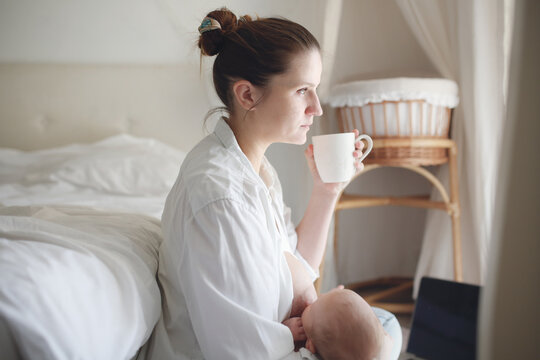 Pensive Tired Cute Caucasian Mom And Newborn, Mom Breastfeeds The Baby By Bed In Light White Bedroom And Drinks Coffee Thoughtfully Near Window. Top View, Toning And Lifestyle In Real Interior