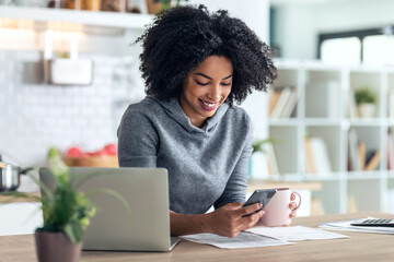 Beautiful afro business woman talking with her mobile phone while working with computer in the kitchen at home.