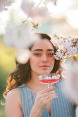 Beautiful young woman with curly hair drinks pink champagne in the blooming sakura garden. Picnic in nature.