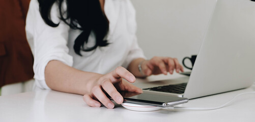 Cropped image of an office woman is charging a smartphone with a wireless charger while typing on a computer laptop.