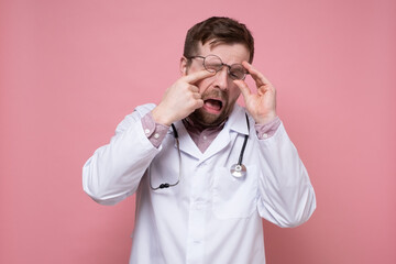 Tired, sleepy doctor with a stethoscope around neck raised glasses and rubs eyes. Pink background.