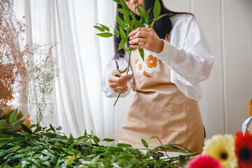 a young brunette woman florist in apron cuts leaves from the stems before starting to create a bouquet in her workshop
