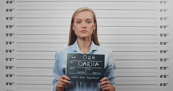 Mugshot of beautiful arrested woman holding sign for photo in police station. Crop view of young female criminal raising head and looking to camera while standing front of metric lineup wall.