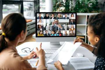 Virtual meeting online, video call. View over shoulders of two women to a computer screen with business leader and successful team, chatting by a video conference, discuss working issues, strategy