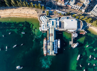 Fototapeta na wymiar Vertical bird's eye aerial evening drone view of Manly wharf, part of the oceanside suburb of Manly, Sydney, New South Wales, Australia. Three ferries docked at the wharf. Manly Cove beach to the left