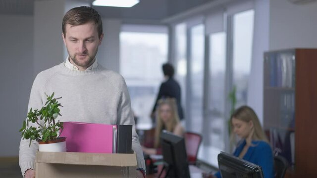 Portrait of jobless frustrated Caucasian man posing with box in office. Sad depressed fired employee looking at camera sighing with blurred colleagues working at background. Crisis and business
