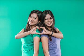 Beautiful twin sisters show a heart from their hands on a green background.