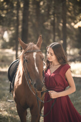 Fototapeta premium Portrait of a smiling young woman hugging her brown horse. A girl in a dress is standing near a horse. The concept of friendship between people and pets.