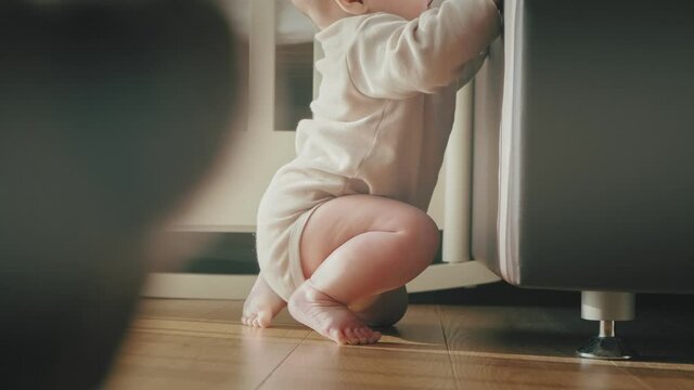 A baby girl in a beige bodysuit slowly gets to her feet holding on to the bed and stands uncertainly