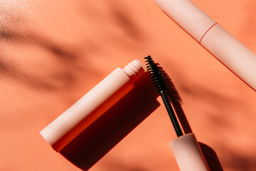 pink clean makeup brush for mascara lies next to an open tube, closed tubes of cosmetics, lip gloss, liquid lipstick, pink eyeliner on a peach background with shadows. Copy space