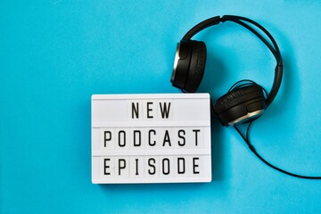 Top view of words NEW PODCAST EPISODE on light box on blue background with headphones. Podcasting...
