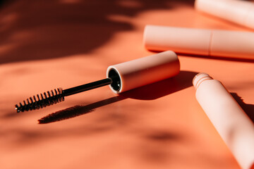 pink clean makeup brush for mascara lies next to an open tube, closed tubes of cosmetics, lip gloss, liquid lipstick, pink eyeliner on a peach background with shadows. Copy space