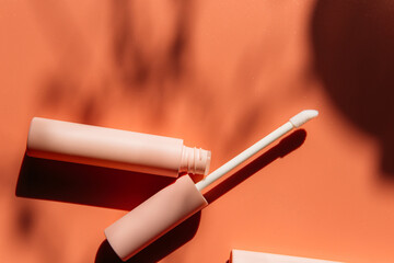 open tube with a brush for liquid lip gloss, liquid lipstick next to closed pink tubes with mascara and eyeliner on a peach background with shadows