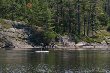 Fototapeta na wymiar Young teenage boy in a life vest paddle boarding on a stand up board at Ontario Provincial Park. Summer recreational activities and water sports. Space for copy. Selective focus.