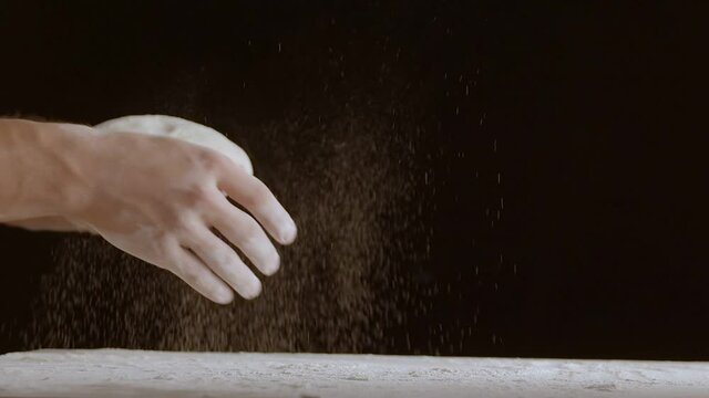 Male hands throw a piece of dough to a wooden table and sprinkle it with flour. Filmed on black background