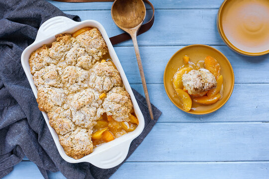 Serving of freshly baked drop biscuit peach cobbler with apron and wooden spoon shot from top view.