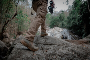 A young man's feet holding a backpack holding binoculars walking on a waterfall background