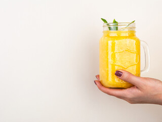 Woman hand holding orange smoothie shake with mint in the glass jar against beige wall.